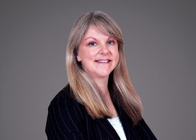 Professional portrait of a person with long blonde hair, wearing a pinstriped black blazer over a light top, smiling against a neutral grey backdrop.