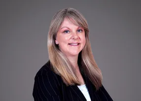 Professional portrait of a person with long blonde hair, wearing a pinstriped black blazer over a light top, smiling against a neutral grey backdrop.