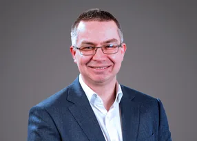 Smiling person in a blue blazer and white shirt, wearing glasses, against a plain grey background.