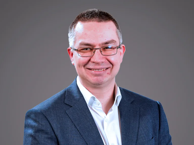 Smiling person in a blue blazer and white shirt, wearing glasses, against a plain grey background.