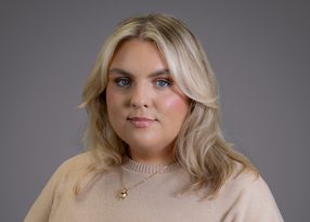 Portrait of a person with shoulder-length blonde hair and blue eyes, wearing a beige sweater and a gold pendant, against a grey studio backdrop.
