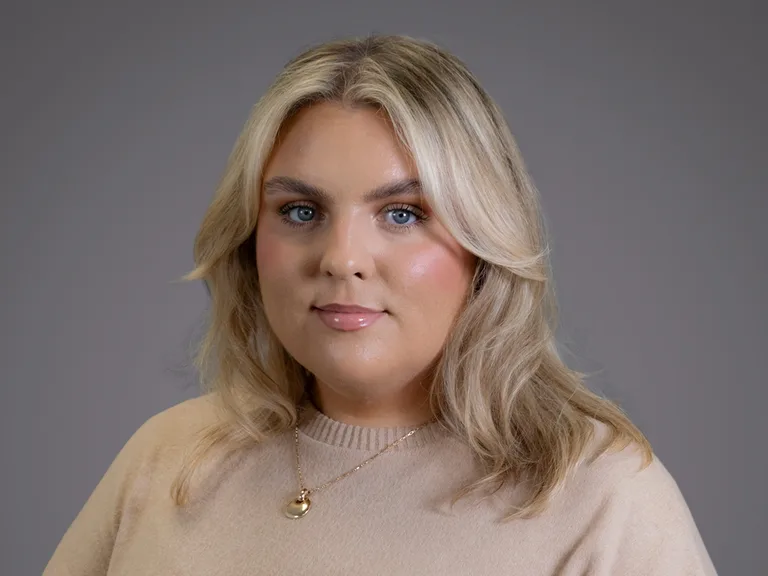 Portrait of a person with shoulder-length blonde hair and blue eyes, wearing a beige sweater and a gold pendant, against a grey studio backdrop.