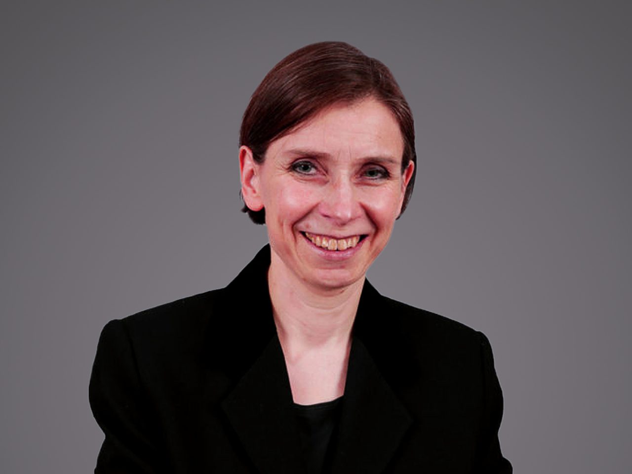 Portrait of a person with short dark hair, wearing a black blazer, smiling against a grey gradient background.