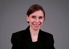 Portrait of a person with short dark hair, wearing a black blazer, smiling against a grey gradient background.