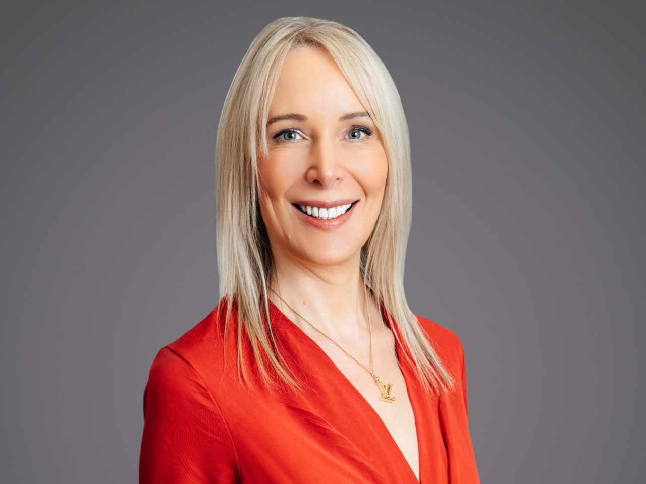Studio portrait of a person with long blonde hair, wearing a black leather jacket, smiling against a neutral grey background.