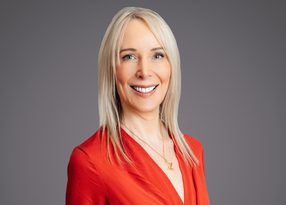 Studio portrait of a person with long blonde hair, wearing a black leather jacket, smiling against a neutral grey background.