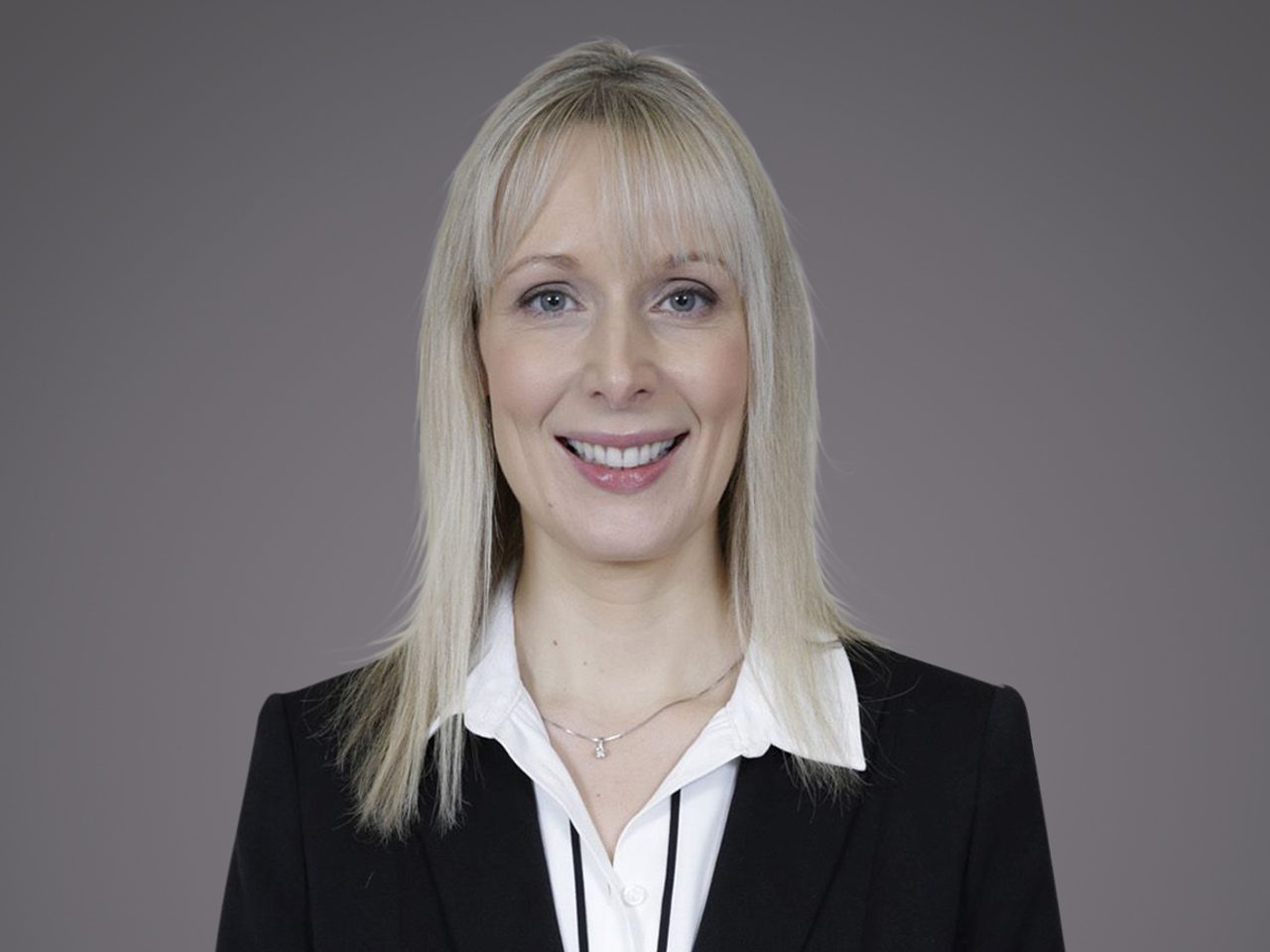 Porträt einer Person mit blonden Haaren, schwarzem Blazer und weißem Hemd, lächelnd vor grauem Hintergrund.