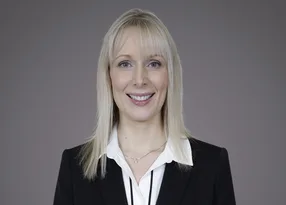 Head-and-shoulders portrait of a blonde individual in a black blazer and white shirt, smiling against a neutral grey studio background.