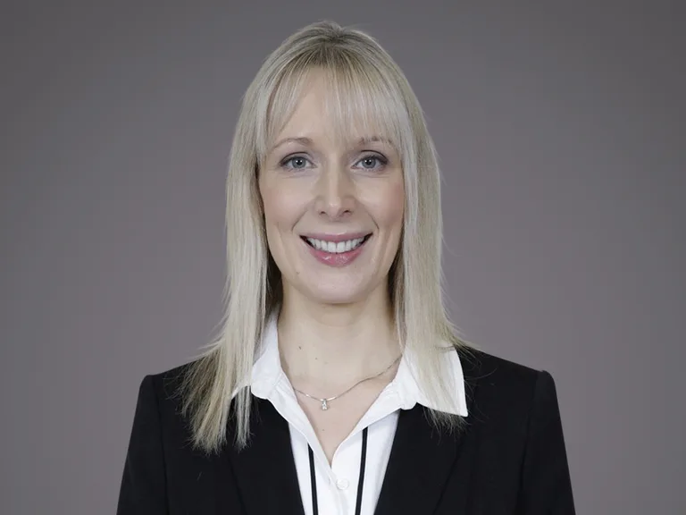 Head-and-shoulders portrait of a blonde individual in a black blazer and white shirt, smiling against a neutral grey studio background.