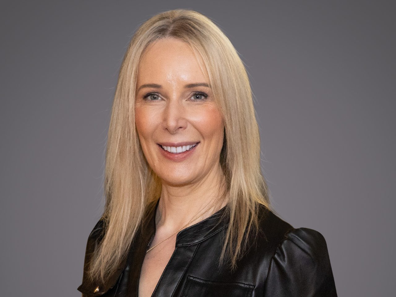 Porträt einer Person mit langem blondem Haar, schwarzer Lederjacke, vor grauem Hintergrund, lächelnd.