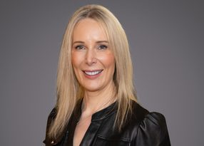 Studio portrait of a person with long blonde hair, wearing a black leather jacket, smiling against a neutral grey background.