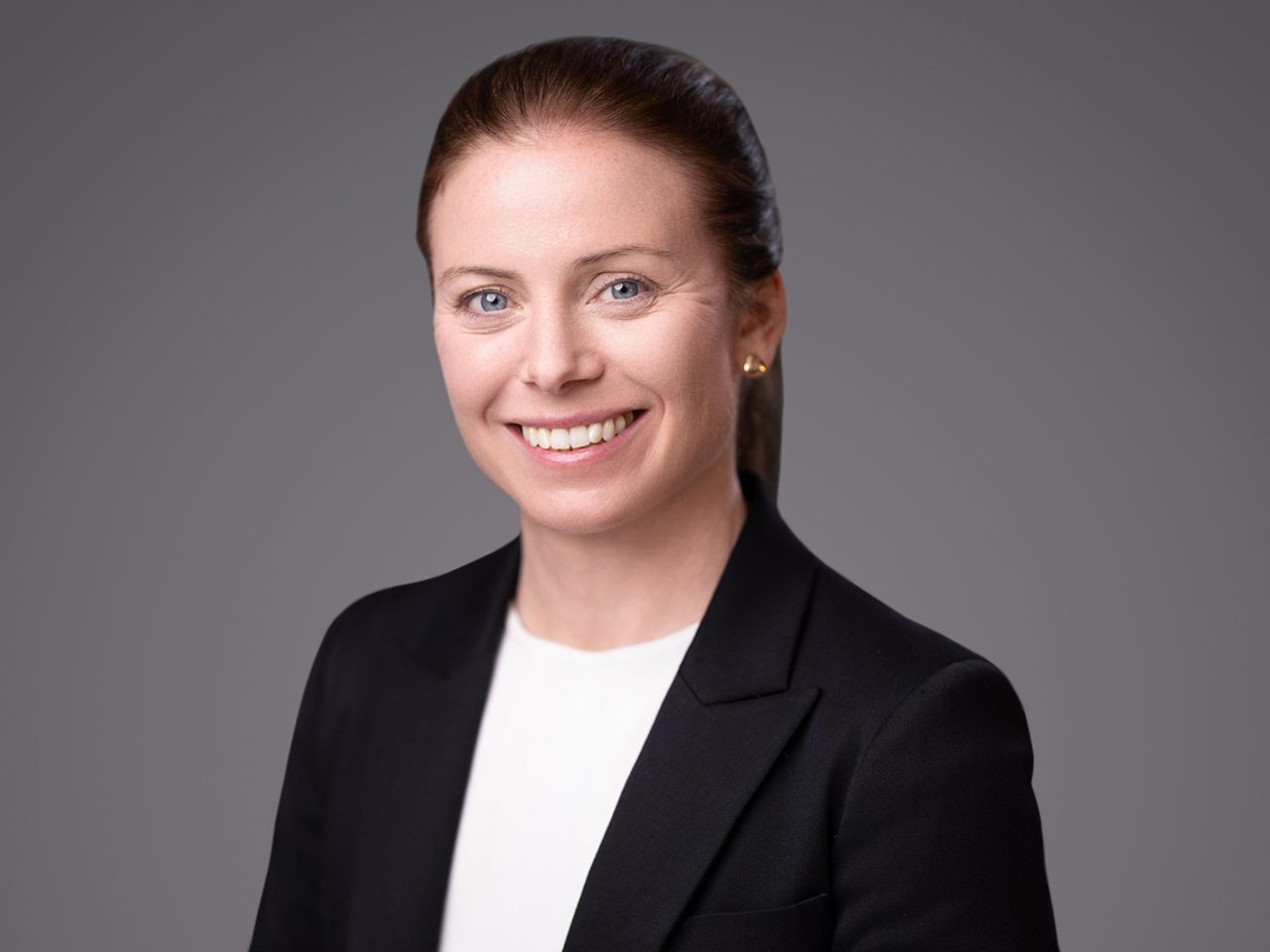 Smiling person with brown hair tied back, wearing a black blazer and white top, against a grey backdrop.
