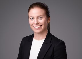 Smiling person with brown hair tied back, wearing a black blazer and white top, against a grey backdrop.