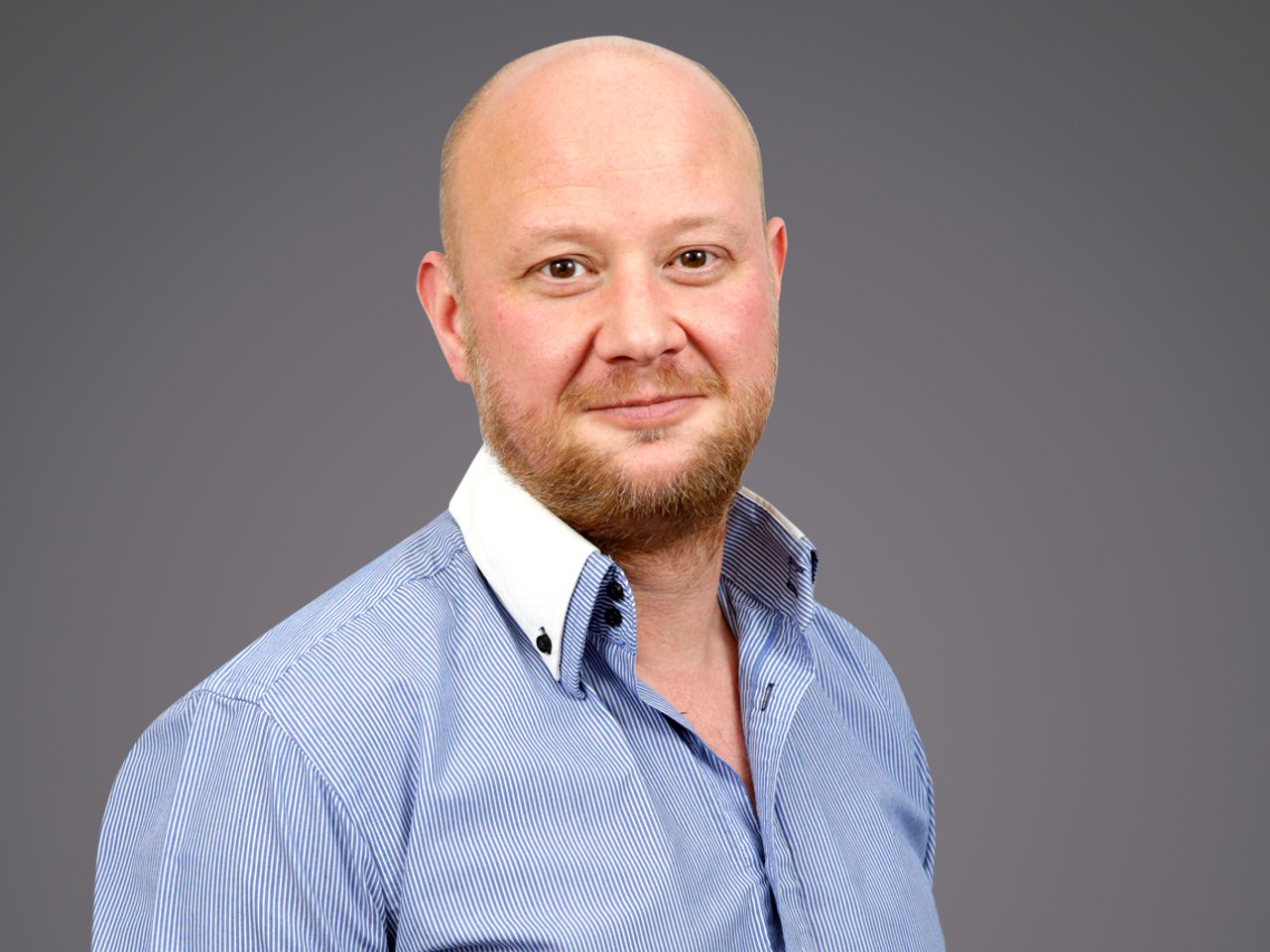 A person with a shaved head and light beard, wearing a blue striped shirt with a white collar, posing against a grey background.