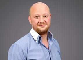 A person with a shaved head and light beard, wearing a blue striped shirt with a white collar, posing against a grey background.