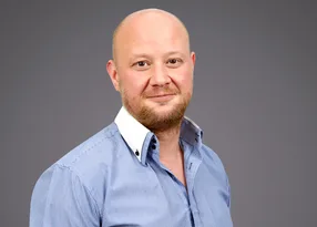 A person with a shaved head and light beard, wearing a blue striped shirt with a white collar, posing against a grey background.