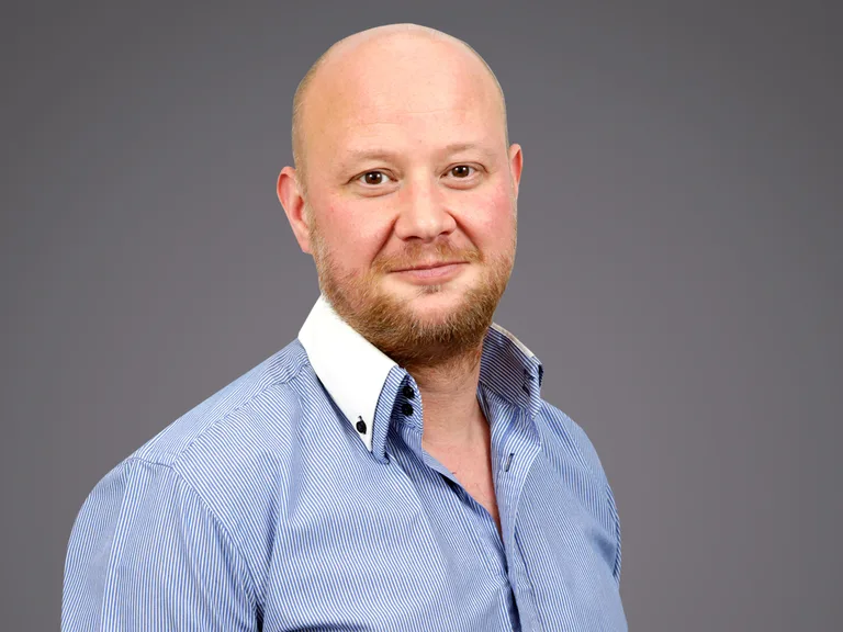 A person with a shaved head and light beard, wearing a blue striped shirt with a white collar, posing against a grey background.