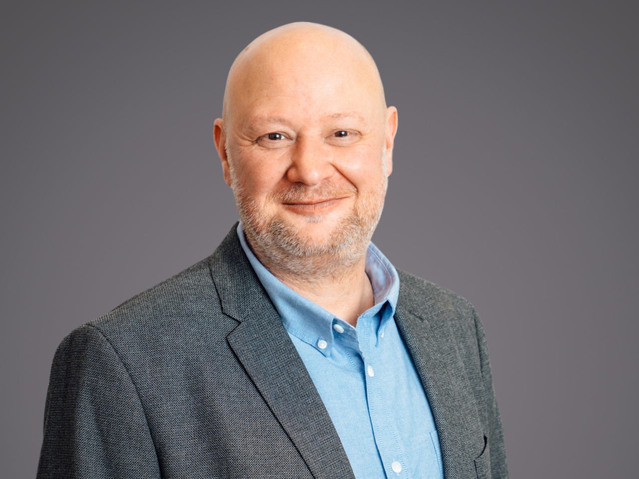 Head-and-shoulders portrait of a bald person with light stubble, wearing a gray blazer and light blue shirt, smiling against a neutral gray backdrop.