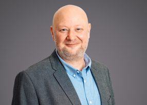Smiling bald individual with a light beard, wearing a grey blazer and light blue shirt, posed against a neutral grey studio background.