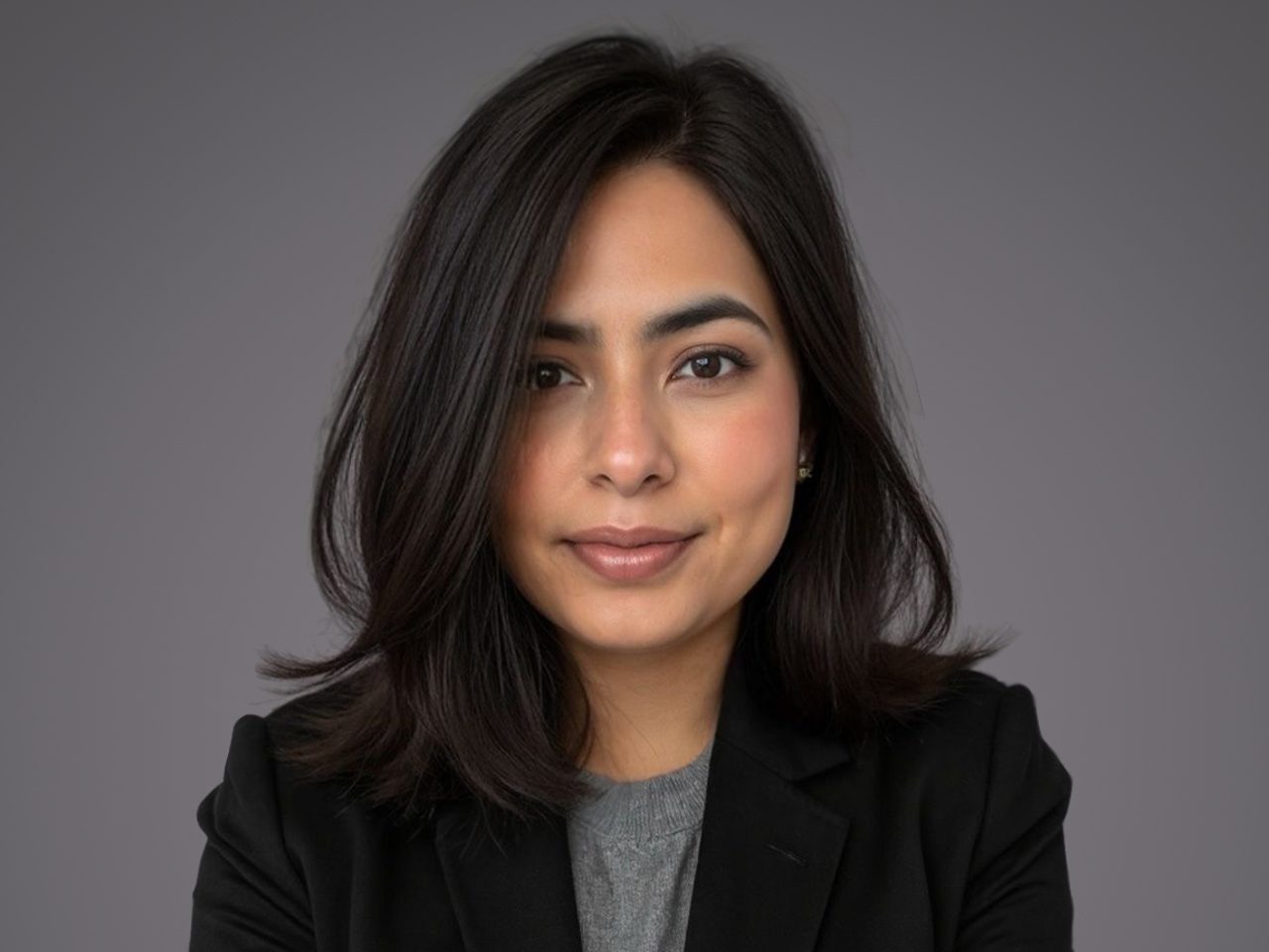 Portrait of a person with shoulder-length dark hair, wearing a grey top and black blazer, smiling softly against a grey studio background.