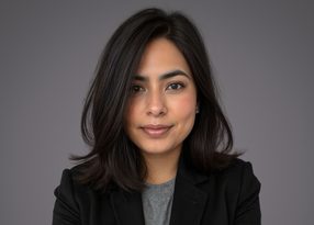 Portrait of a person with shoulder-length dark hair, wearing a grey top and black blazer, smiling softly against a grey studio background.