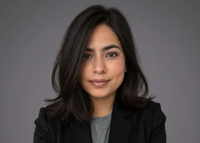 Portrait of a person with shoulder-length dark hair, wearing a grey top and black blazer, smiling softly against a grey studio background.