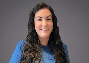 Portrait of a person with long dark wavy hair, wearing a blue top, smiling against a neutral grey backdrop.
