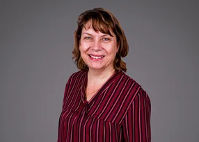 A person with short brown hair wearing a maroon striped blouse, smiling warmly against a plain grey studio background.