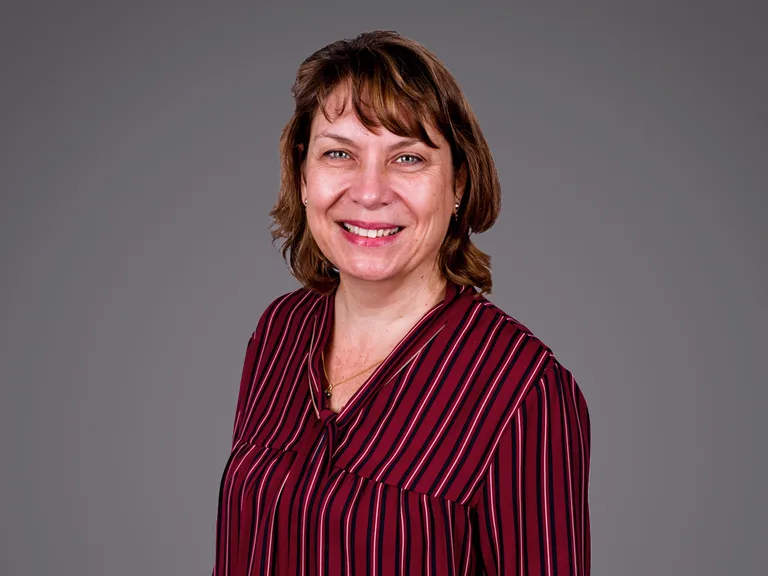 Smiling person with short brown hair in a maroon striped blouse, posing against a neutral gray background.