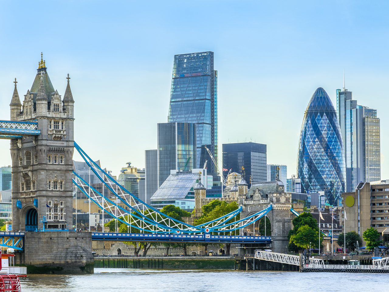 The Tower Bridge spans the River Thames, with modern skyscrapers, including The Gherkin and Cheesegrater, in the background under a clear blue sky.