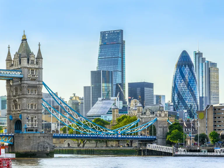 The Tower Bridge spans the River Thames, with modern skyscrapers, including The Gherkin and Cheesegrater, in the background under a clear blue sky.