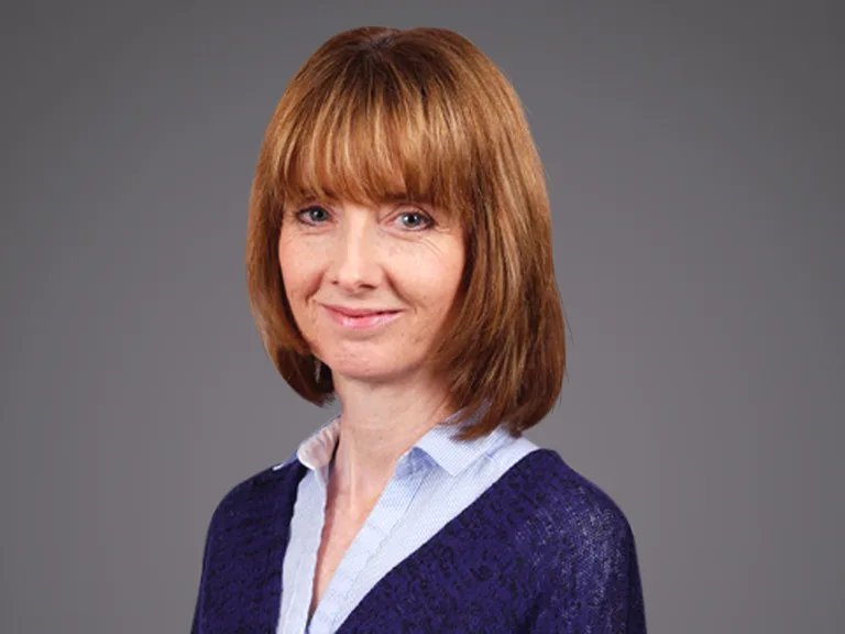 Person with shoulder-length light brown hair and bangs, wearing a blue textured cardigan over a pale blue shirt, smiling against a grey studio background.