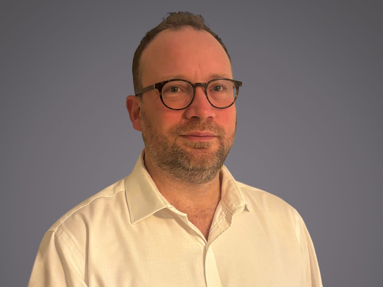 Portrait of a person with short hair, a beard, and round glasses, wearing a light button-down shirt, against a plain gray-blue background.
