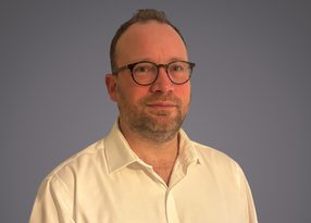 Portrait of a person with short hair, a beard, and round glasses, wearing a light button-down shirt, against a plain gray-blue background.