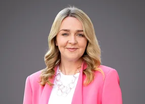Portrait of a person with blonde hair, wearing a pink blazer over a white top and a geometric necklace, against a grey background.