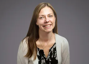 Smiling person with long light brown hair, wearing a black floral blouse and beige cardigan, against a plain grey backdrop.