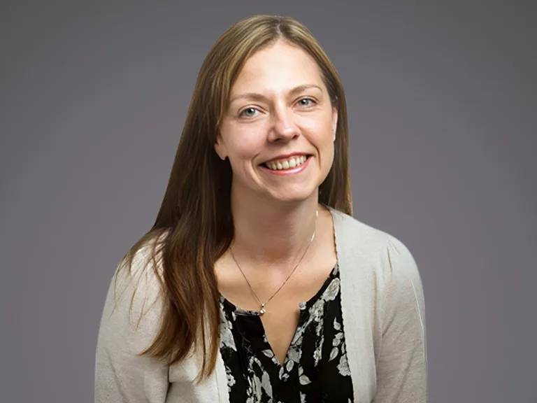 Portrait of a person with long light brown hair, smiling, wearing a beige cardigan over a black floral blouse, against a gray gradient background.