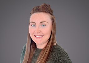 Portrait of a person with long brown hair styled up, blue eyes, and a friendly smile, wearing a green patterned sweater against a gray backdrop.
