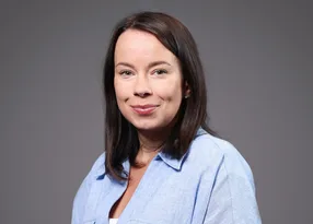 Person with shoulder-length dark hair, wearing a light blue shirt, smiling softly at the camera against a neutral grey background.