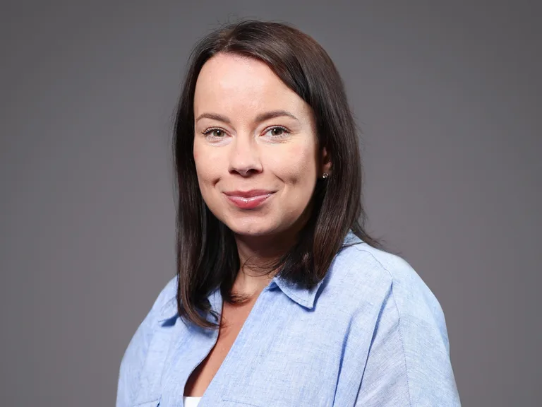 Portrait of a person with shoulder-length dark hair, wearing a light blue collared shirt, smiling softly against a gray background.