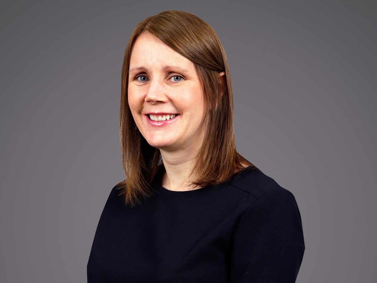 A person with shoulder-length brown hair wearing a dark top, smiling against a plain grey background.