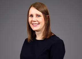 A person with shoulder-length brown hair wearing a dark top, smiling against a plain grey background.
