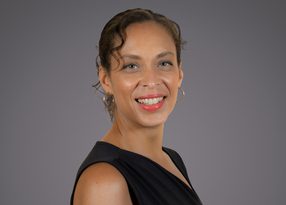 Smiling person with short, wavy hair wearing hoop earrings and a black sleeveless top, posed against a gray studio background.