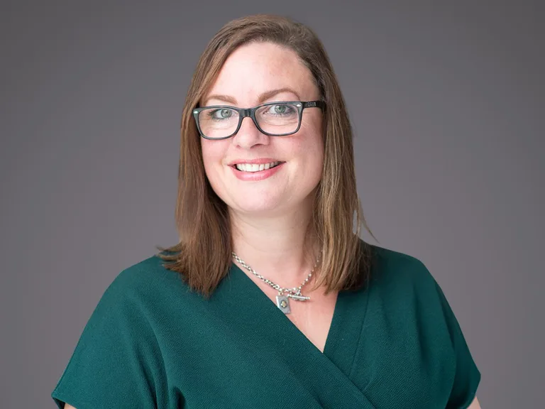 A person with shoulder-length light brown hair, black-framed glasses, and a teal green V-neck top, smiling against a plain grey backdrop.