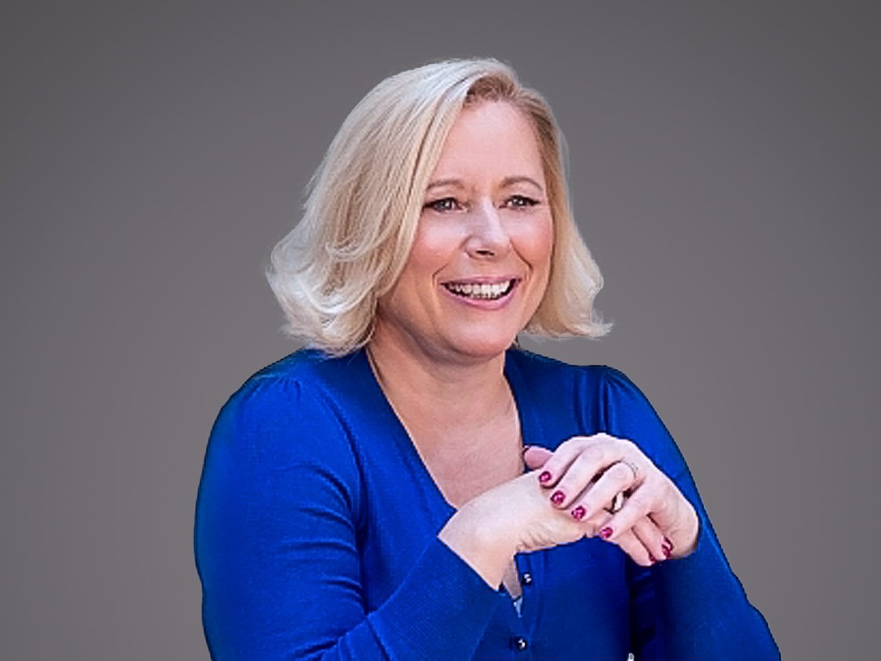 A smiling person with shoulder-length blond hair wearing a blue long-sleeve top, hands clasped in front against a grey gradient background.