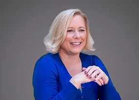 A smiling person with shoulder-length blond hair wearing a blue long-sleeve top, hands clasped in front against a grey gradient background.
