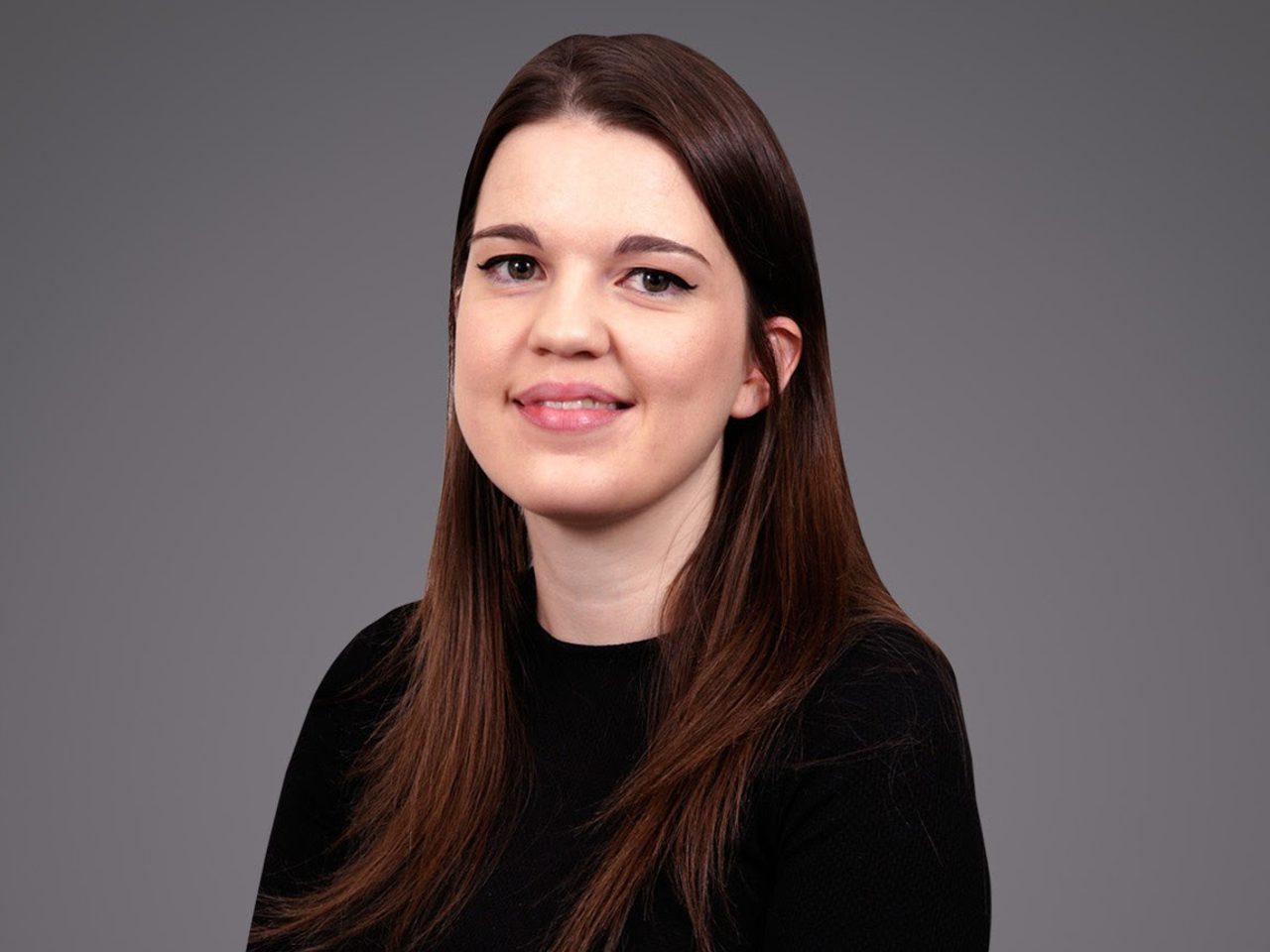 Portrait of a person with long brown hair, a subtle smile, wearing a black top, against a gray studio backdrop.