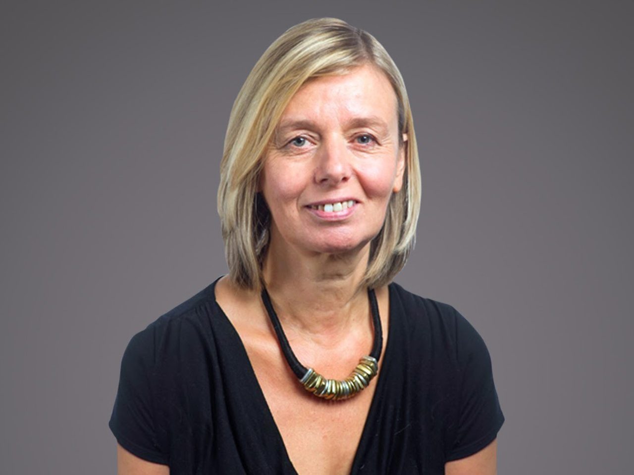 Smiling person with shoulder-length blonde hair, wearing a black top and a chunky beaded necklace, against a neutral grey background.