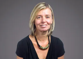 Smiling person with shoulder-length blonde hair, wearing a black top and a chunky beaded necklace, against a neutral grey background.