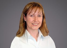 Person with shoulder-length light brown hair wearing a white button-up shirt, smiling against a neutral gray gradient background.